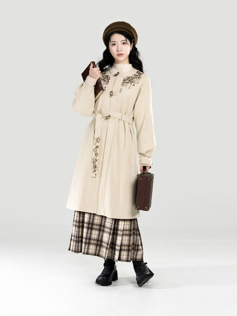 Woman in a beige coat with floral embroidery and plaid skirt holding a brown bag on a white background