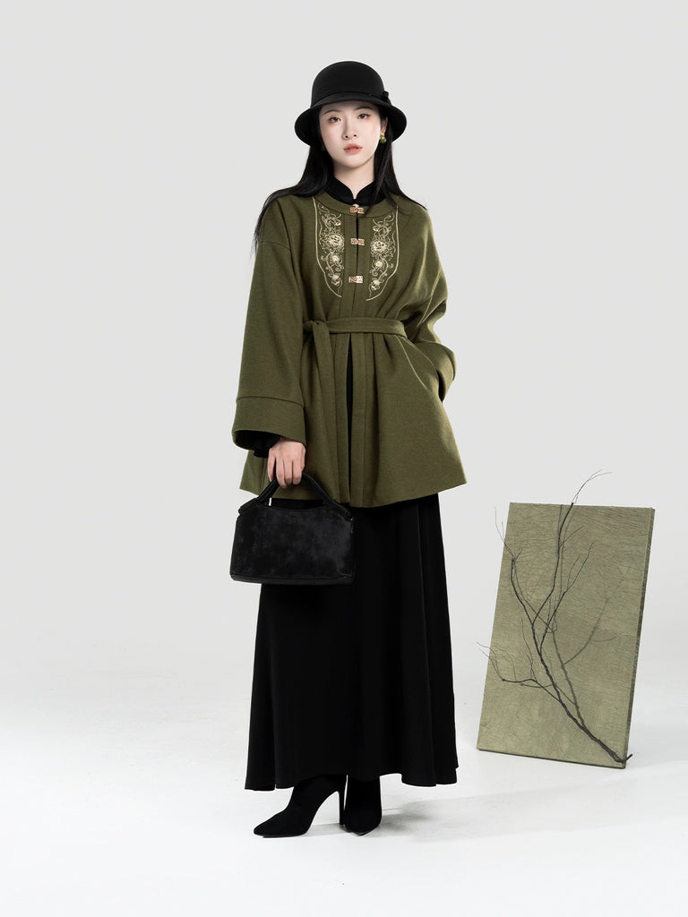 Woman wearing a green embroidered coat and black hat, holding a black bag, with a white background.